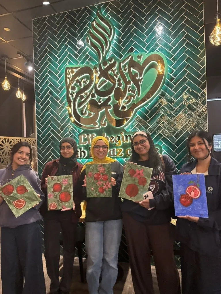 Five guests holding up their finished pomegranate canvases in front of a teal cafe wall.