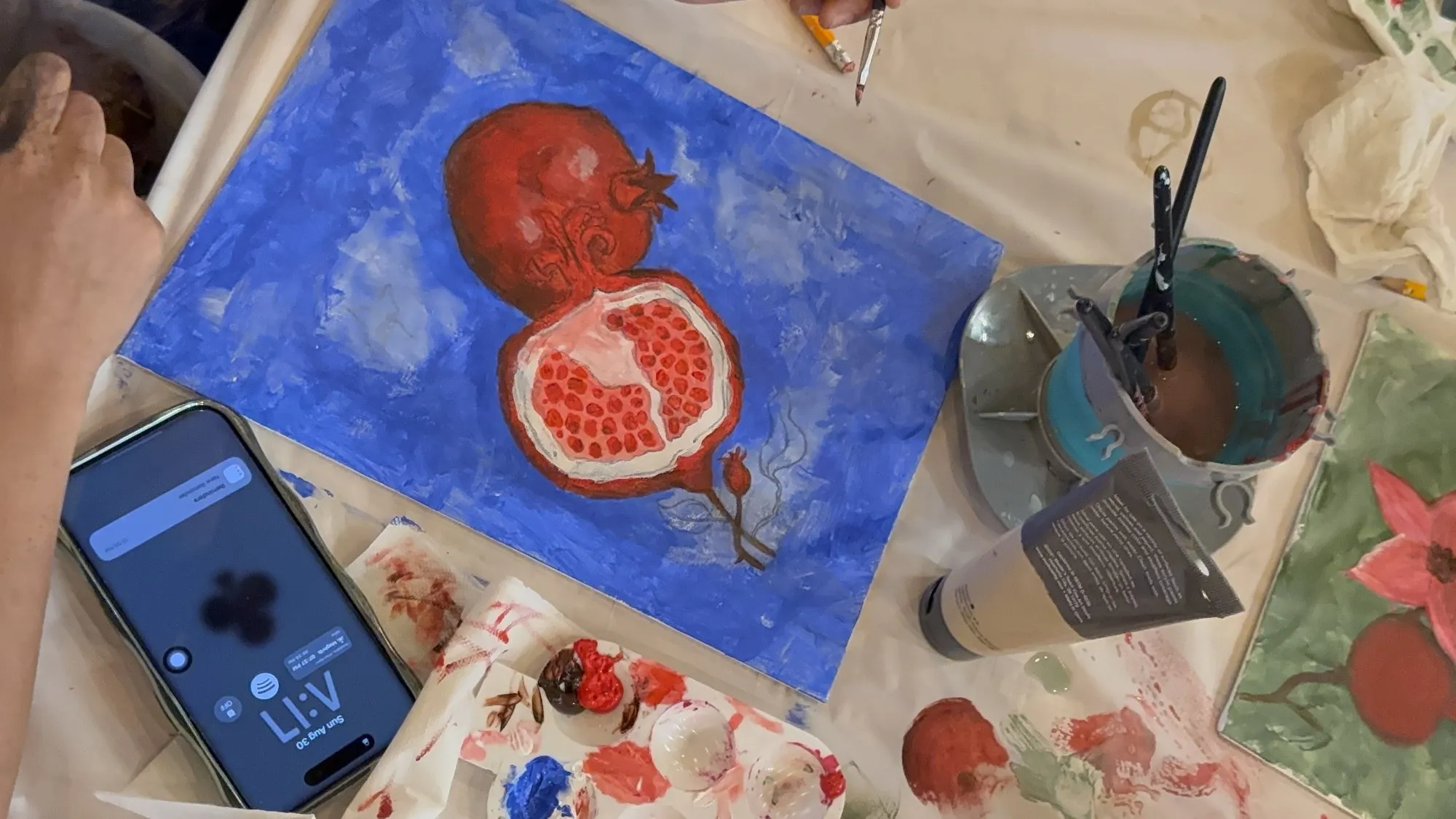 A pomegranate painting in progress on a blue canvas, beside a palette of reds.