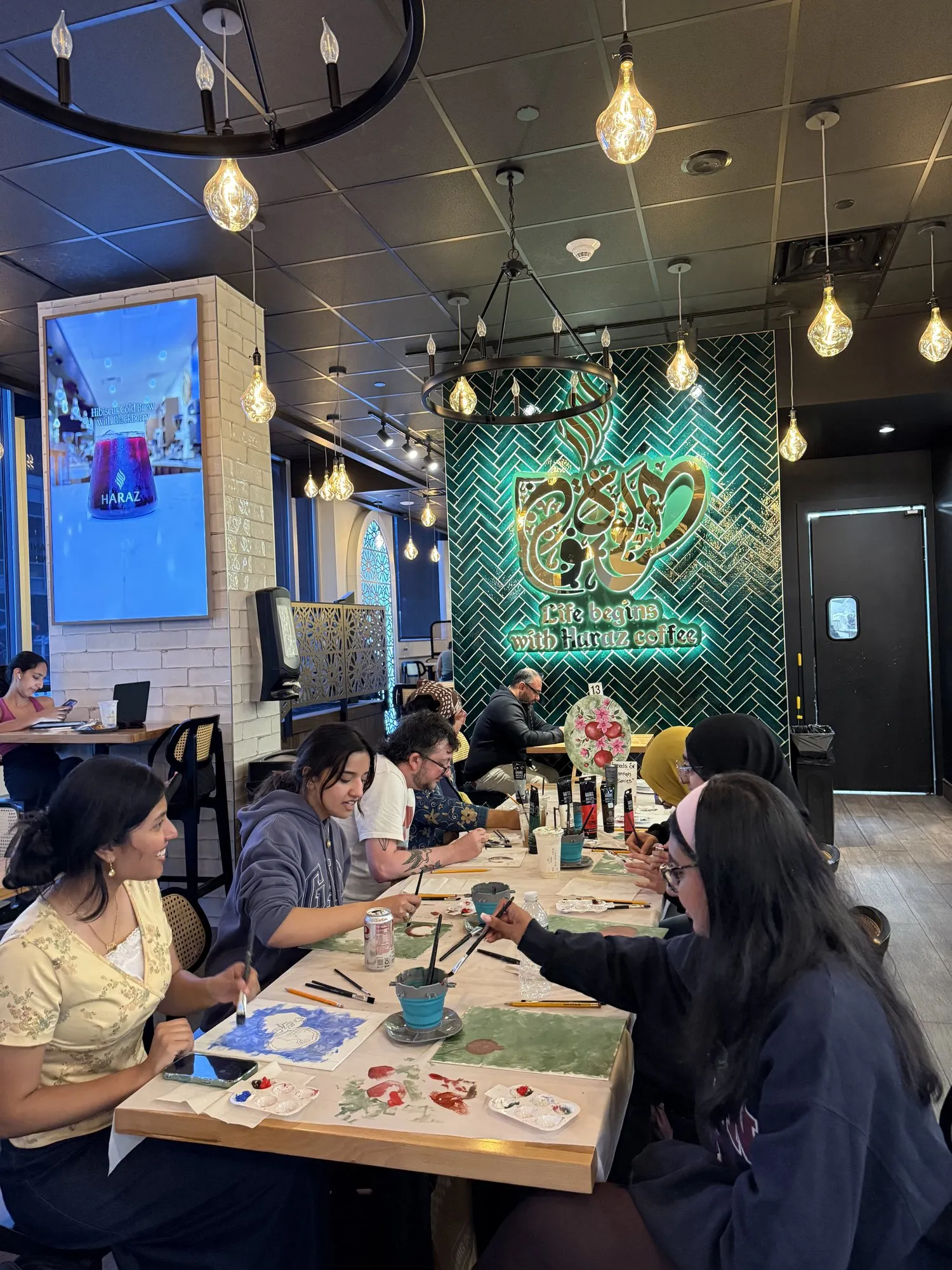 A wide view of a cafe under pendant lights, guests painting at every table.