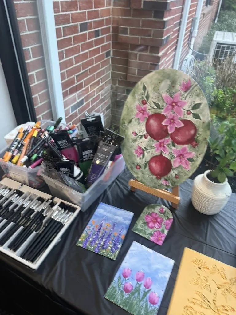 Finished works displayed on a table by a window, including a painted pomegranate panel.