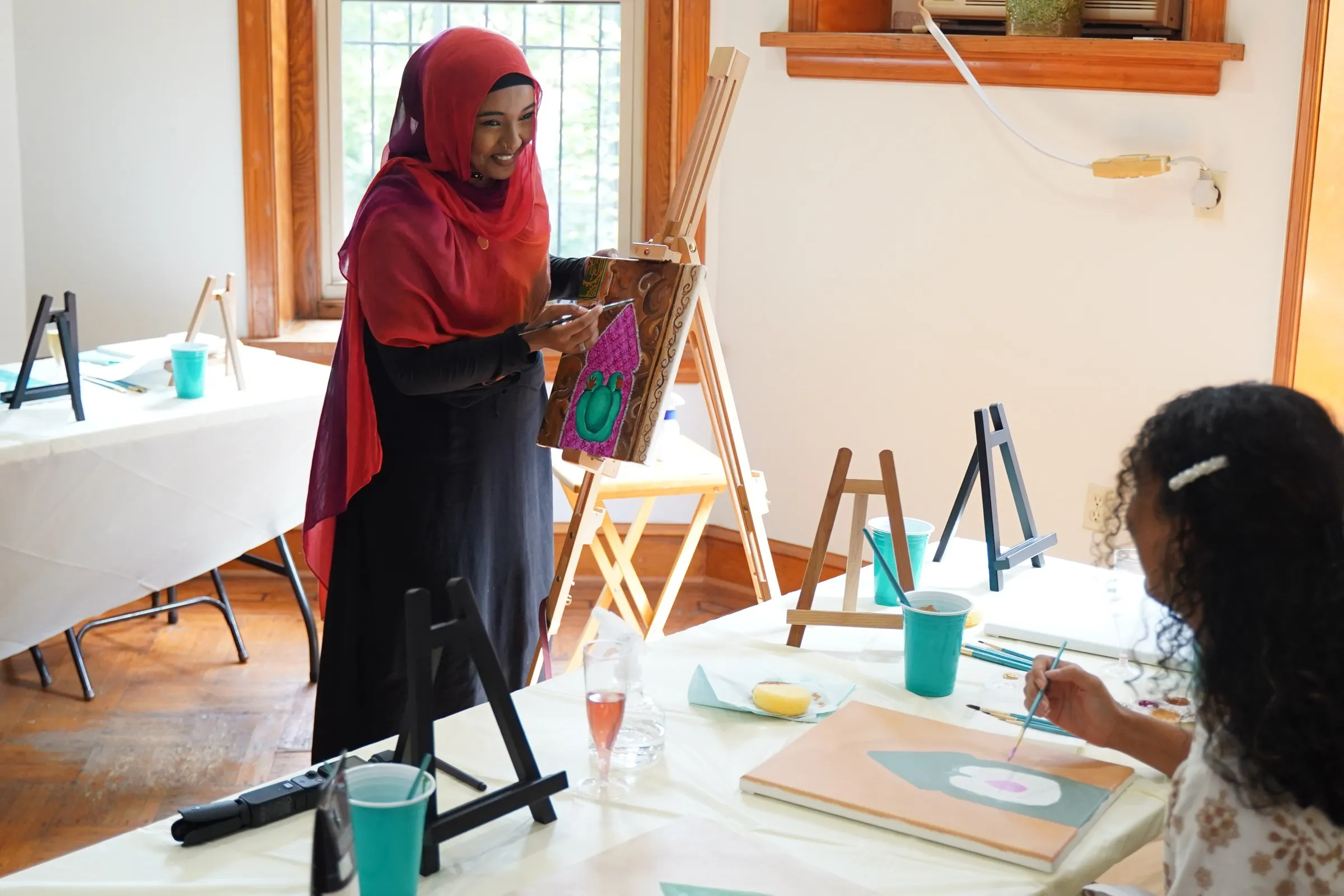 Hanan, in a red headscarf, holding up a painting as she leads a class in a sunlit studio lined with easels.